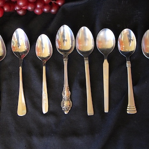 May include: An assortment of silver spoons with various handle designs, arranged on a black surface. The spoons have different styles, including floral and geometric patterns. Red grapes are in the upper left corner.