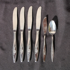May include: A set of six silver-colored utensils, including five knives, a butter knife, and a spoon, arranged on a dark surface. The knife handles feature a rose design. A bunch of red grapes is in the upper right corner.