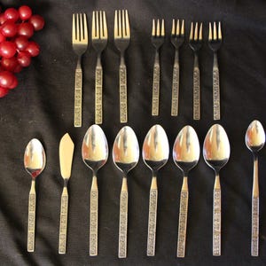 May include: A collection of vintage silverware, including forks, spoons, and a butter knife, arranged on a black surface. The silverware has a decorative pattern on the handles. A cluster of red grapes is in the upper left corner.
