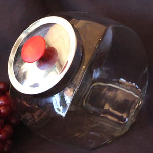 May include: A clear glass canister with a slanted opening and a circular metal lid featuring a bright red knob handle. The canister is positioned next to a cluster of dark red grapes, showcasing its transparent body and reflective metallic lid.