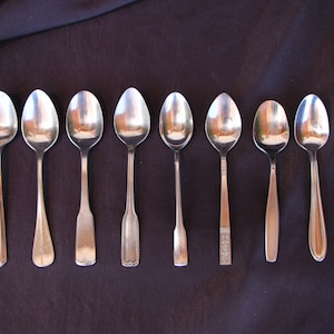 May include: Eight silver spoons arranged in a row on a dark purple surface. The spoons are all different styles and sizes.