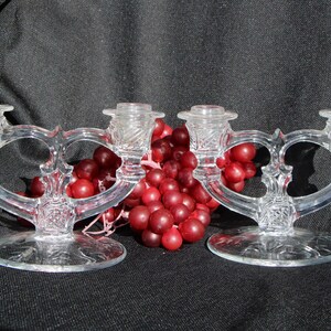 May include: Two clear glass candle holders with a heart-shaped design, each holding two candle cups. The candle holders are on a round base. A bunch of red grapes is between the candle holders.