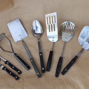 May include: A set of seven kitchen utensils with black handles. The utensils include a potato masher, a pizza cutter, a spatula, a slotted spoon, a slotted turner, a whisk, and a large slotted spatula.