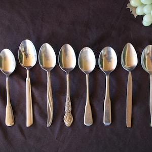 Puede incluir: Ocho cucharas de plata de diferentes estilos y tamaños están alineadas sobre una superficie marrón. Las cucharas son todas brillantes y reflejan la luz.