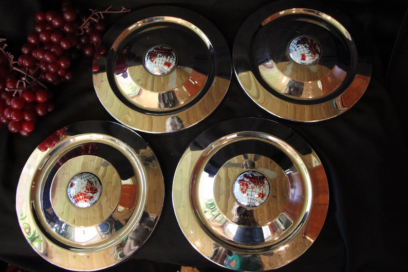 Puede incluir: Cuatro platos de metal vintage con una superficie brillante y reflectante. Cada plato presenta un dise&ntilde;o de globo central con continentes rojos y blancos. Los platos tienen un centro negro y un borde dorado.