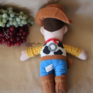 Puede incluir: Un juguete de peluche de Woody de Toy Story. Lleva su caracter&iacute;stico sombrero de vaquero marr&oacute;n, una camisa de cuadros amarillos y marrones, pantalones vaqueros azules y un cintur&oacute;n marr&oacute;n con una hebilla plateada. Tiene un chaleco marr&oacute;n con un patr&oacute;n de vaca blanco y negro y una bandana roja alrededor del cuello.