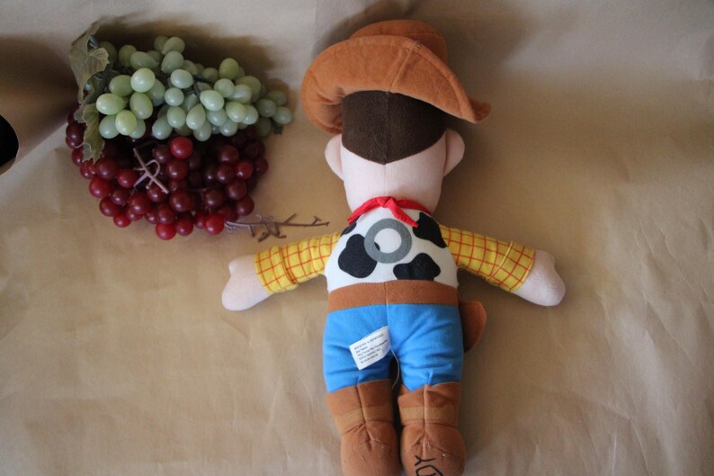 Puede incluir: Un juguete de peluche de Woody de Toy Story. Lleva su caracter&iacute;stico sombrero de vaquero marr&oacute;n, una camisa de cuadros amarillos y marrones, pantalones vaqueros azules y un cintur&oacute;n marr&oacute;n con una hebilla plateada. Tiene un chaleco marr&oacute;n con un patr&oacute;n de vaca blanco y negro y una bandana roja alrededor del cuello.