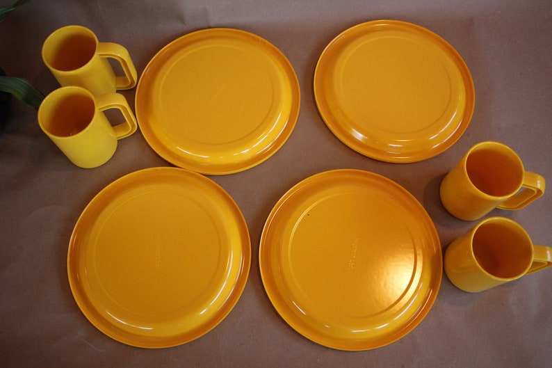 Op de afbeelding: Een set van vier gele plastic borden en vier gele plastic bekers. De borden zijn rond en hebben een licht verhoogde rand. De bekers hebben een handvat en zijn iets hoger dan de borden.