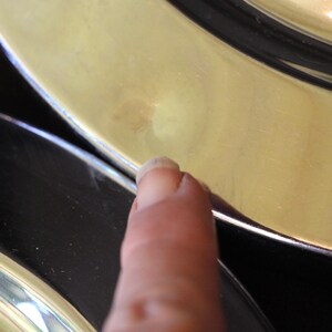 Puede incluir: Primer plano de un dedo tocando el borde de un plato dorado. El plato tiene un borde negro y una superficie brillante. La imagen est&aacute; bien iluminada, destacando las texturas y los colores del plato.