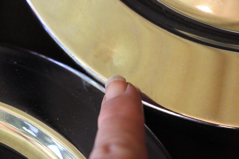 Puede incluir: Primer plano de un dedo tocando el borde de un plato dorado. El plato tiene un borde negro y una superficie brillante. La imagen est&aacute; bien iluminada, destacando las texturas y los colores del plato.