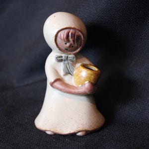 May include: A ceramic figurine of a girl wearing a white hooded dress and holding a small brown pot. The girl has brown hair and a blue ribbon around her neck.