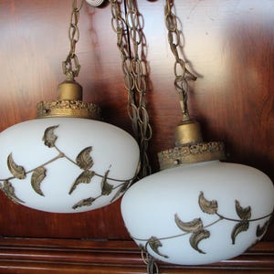 May include: Two vintage pendant lights with white glass shades and brass-colored accents. Each shade features a decorative leaf design. The lights hang from brass chains, showcasing an antique aesthetic.
