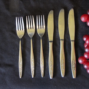 May include: A set of six vintage silverware pieces, including three forks and three knives, arranged on a dark surface. The silverware has a floral design on the handles. A cluster of red grapes is in the background.