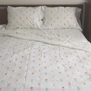 May include: White bed linens with a repeating pattern of small, colorful tulips. The set includes a flat sheet, fitted sheet, and two pillowcases. The bedding is on a bed with a wooden headboard and a wooden dresser.