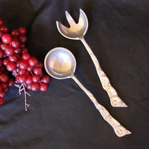 May include: A set of two silver serving utensils, a fork and a spoon, with ornate handles. The utensils are on a black fabric background.