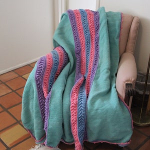 May include: A teal crocheted throw blanket with pink, purple, and blue stripes. The blanket is draped over a pink armchair.