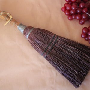 May include: A small hand broom with a natural wooden handle, a light-colored leather hanging loop, and a silver-toned metal ferrule securing dark brown bristles that are bound together with two horizontal rows of black thread.