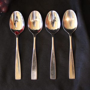 May include: Four silver spoons with oval bowls and tapered handles are arranged on a black surface. The spoons reflect the surrounding environment. A cluster of red grapes is visible in the upper left corner.