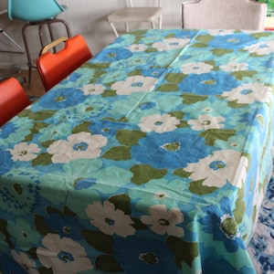 May include: A rectangular tablecloth with a floral pattern in shades of blue, white, and green covers a table. The tablecloth is the focal point, with orange chairs and bar stools in the background.