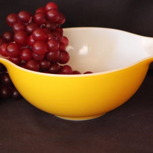 May include: A bright yellow mixing bowl with a white interior and a pouring spout on one side. The bowl is filled with a large cluster of deep red grapes, which spill over the edge of the bowl onto the surface below the container.