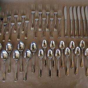 May include: A set of silverware, including forks, spoons, and knives, laid out on a brown surface. The silverware is made of polished stainless steel.