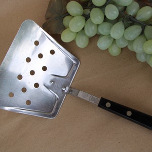 May include: A silver slotted spatula with a black handle. The spatula has a wide, flat head with holes for draining liquids.