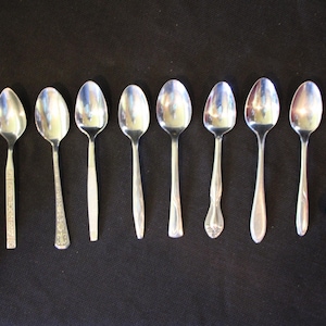 Puede incluir: Ocho cucharaditas de plata con diseños diferentes sobre un fondo negro.