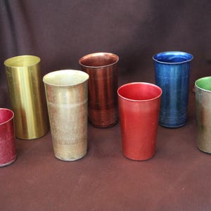 May include: A collection of seven cylindrical metal tumblers in various colors including red, gold, copper, blue, and green. Each tumbler features a smooth or ribbed texture along the base, with varying heights and metallic finishes throughout.
