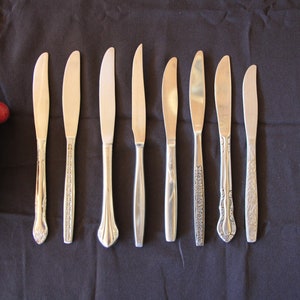 May include: Eight silver butter knives with different designs on a black fabric background.