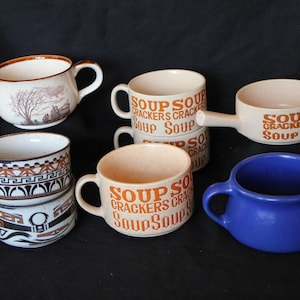May include: A collection of ceramic soup mugs and bowls in various designs and colors. Includes a blue mug, mugs with landscape and geometric patterns, and mugs with "SOUP CRACKERS SOUP" text. The mugs have handles.