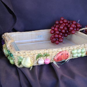 May include: A woven basket with a glass baking dish inside. The basket is decorated with colorful fruit and vegetable designs. A bunch of red grapes sits on top of the basket.