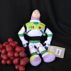 May include: A green, white, and purple plush toy of Buzz Lightyear from the Toy Story franchise. The toy is sitting on a black surface with a bunch of red grapes.