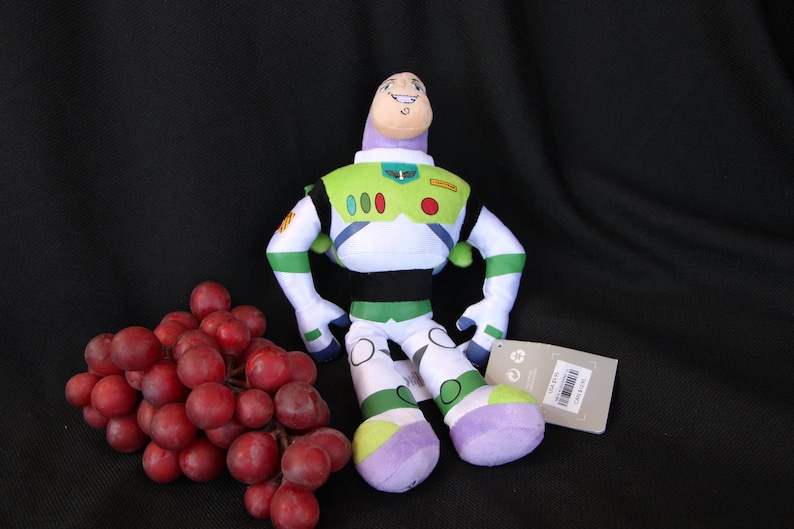 May include: A green, white, and purple plush toy of Buzz Lightyear from the Toy Story franchise. The toy is sitting on a black surface with a bunch of red grapes.