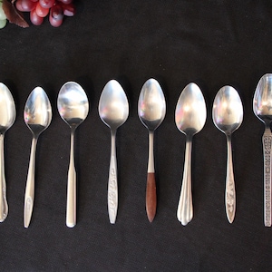 May include: Eight silver teaspoons with varying designs on the handles. The spoons are arranged in a row on a black surface.
