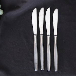 May include: Four silver dinner knives with a modern design, laid out on a black surface.
