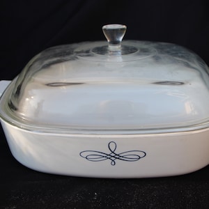 May include: White casserole dish with a clear glass lid and a clear glass knob. The dish has a decorative blue swirl design on the front and white handles on each side. The dish is on a black surface.