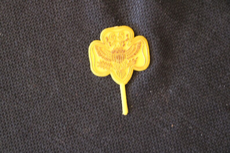 May include: A yellow plastic Girl Scout pin in the shape of a four-leaf clover with a stylized eagle design in the center.