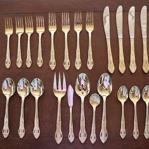 May include: A set of gold-plated silverware, including forks, spoons, and knives, arranged on a wooden surface. The silverware has an ornate design and is in excellent condition.