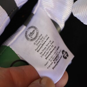 May include: White tag with black text that reads "Disney Pixar", "Disney Store", "DisneyStore.com", "Pasadena, CA 91105", "The Disney Store Ltd", "3 Queen Caroline Street", "London W6 9PE", "DisneyStore.eu", "〒105-6355 東京都", "ウォルト・ディズニー・ジャパン", "ディズニー・ストア", "虎ノ門ヒルズ", "DisneyStore.com".