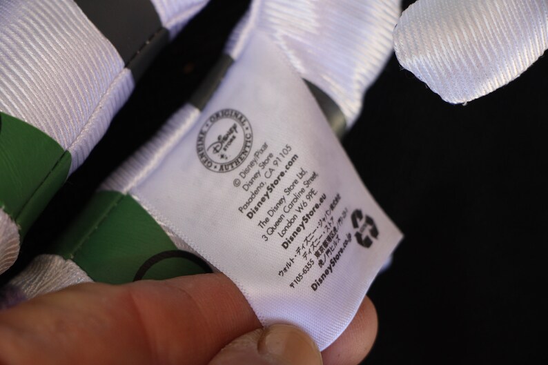May include: White tag with black text that reads "Disney Pixar", "Disney Store", "DisneyStore.com", "Pasadena, CA 91105", "The Disney Store Ltd", "3 Queen Caroline Street", "London W6 9PE", "DisneyStore.eu", "〒105-6355 東京都", "ウォルト・ディズニー・ジャパン", "ディズニー・ストア", "虎ノ門ヒルズ", "DisneyStore.com".