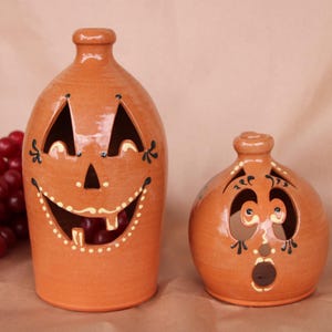 May include: Two ceramic pumpkin-shaped candle holders with painted faces. The larger holder is orange with a wide, toothy grin and black eyes. The smaller holder is a similar orange color with a rounder face and black eyes.