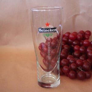 May include: A clear glass beer tumbler featuring the Heineken logo in black text with a red star above it. The glass has a tapered cylindrical shape with a thick base. The brand name is printed horizontally near the top and vertically below.