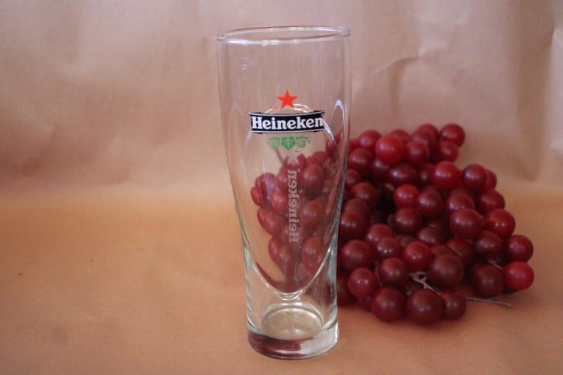 May include: A clear glass beer tumbler featuring the Heineken logo in black text with a red star above it. The glass has a tapered cylindrical shape with a thick base. The brand name is printed horizontally near the top and vertically below.