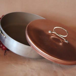May include: A round, shallow cooking pot with a brushed silver-toned metal exterior and a smooth, copper-colored lid. The pot features two silver-toned metal loop handles on the sides, and the lid has a matching silver-toned metal loop handle.