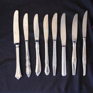 May include: A set of eight silver butter knives with ornate handles. The knives are arranged in a row on a black surface.