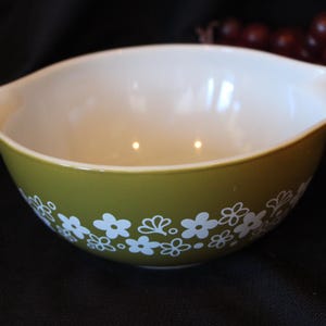 May include: An olive green Pyrex mixing bowl with a white floral pattern around the base. The interior is white, and the bowl has two handles. A classic kitchenware item.