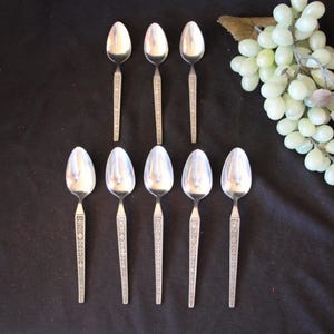 May include: Set of ten silver spoons with an ornate design. The spoons are arranged in two rows of five on a black surface.