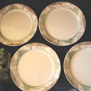 May include: Four off-white ceramic plates with a decorative border featuring green and brown leaf patterns. A bunch of green grapes with leaves is visible on the left side of the image. The plates are arranged on a dark surface.