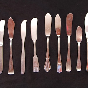 May include: A collection of nine butter knives with various blade shapes and handle designs. The knives are made of silver-colored metal and are arranged on a dark surface. Some blades are curved, while others are straight.