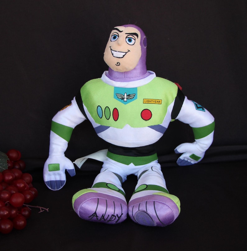 May include: A plush toy of Buzz Lightyear from the Toy Story franchise. The toy is wearing his signature green and white space ranger suit with a purple helmet. The toy has a big smile and is sitting with his legs crossed. The toy's feet are purple and have the name "ANDY" written on them.